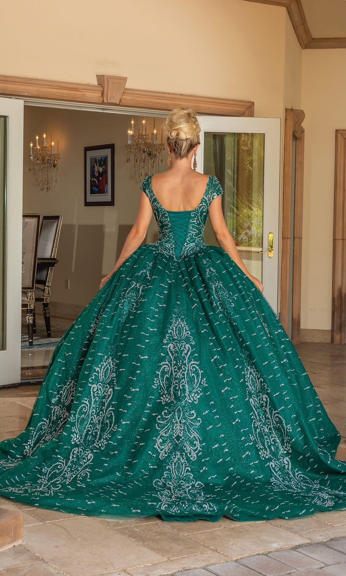A woman in the Quinceanera Dress 1805 By Dancing Queen, featuring hunter green color, cap sleeves, and a sweetheart neckline, stands facing away outside open double doors to a bright formal room.